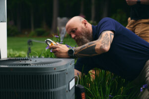 A bearded man with tattoos uses a tool to inspect an outdoor air conditioning unit surrounded by plants.