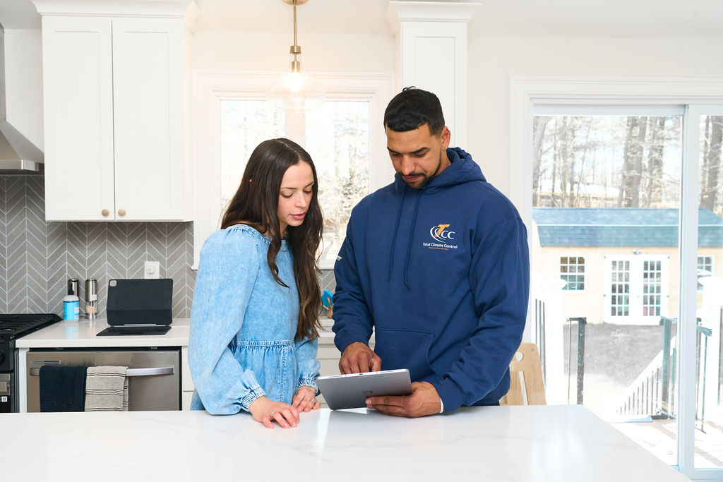 Two people stand in a bright kitchen, looking at a tablet on the counter. One person wears a blue dress, and the other wears a blue hoodie with a logo.