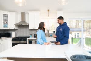 A woman and a man stand talking in a bright, modern kitchen. The man holds a tablet, and both appear engaged in conversation about water line services NH. Large windows show a view of a yard outside.