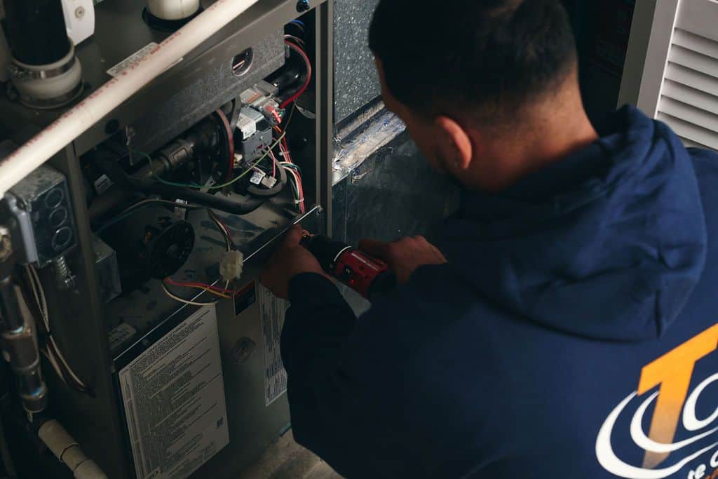 A person in a blue hoodie uses a power drill to repair or install components inside a heating unit